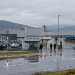 Automobilový závod v Bratislave za daždivého rána; priemyselné haly, nakladacie rampy a robotníci zozadu.