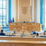 Široký záber rokovacej sály Národnej rady SR, lavice, mikrofóny a vzdialené postavy odzadu v dennom svetle.