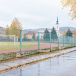 Staršie športovisko za plotom pri ceste v slovenskom meste, zamračené popoludnie, postava v diaľke odchádza.