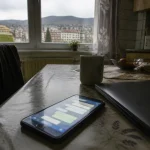 Smartfón na kuchynskom stole so slabým displejom a abstraktnými chatovými bublinami, vedľa laptop, denné svetlo.